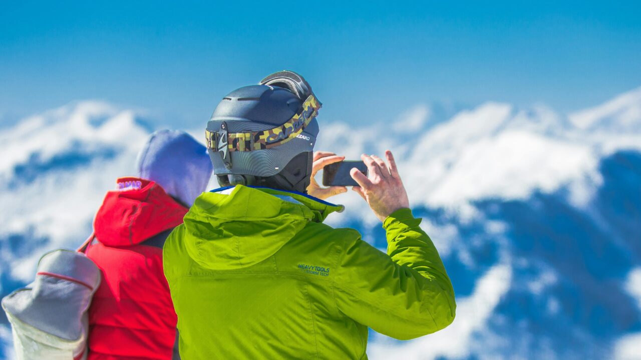 Man in Green Jacket and Gray Helmet Holding Phone Standing Next to Person in Red and Black Jacket