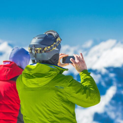 Man in Green Jacket and Gray Helmet Holding Phone Standing Next to Person in Red and Black Jacket