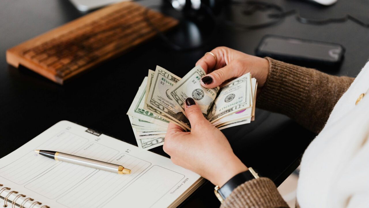 Unrecognizable elegant female in sweater counting dollar bills while sitting at wooden table with planner and pen