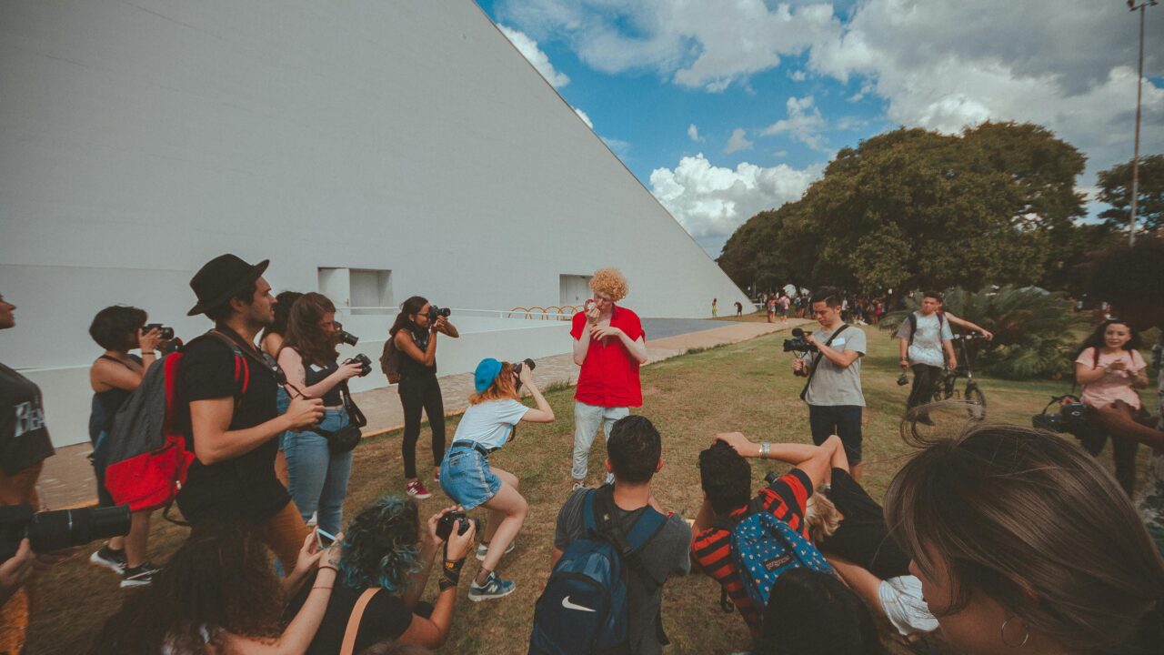 A group of people gathered outdoors, photographing a person in a red shirt, against a geometric backdrop.