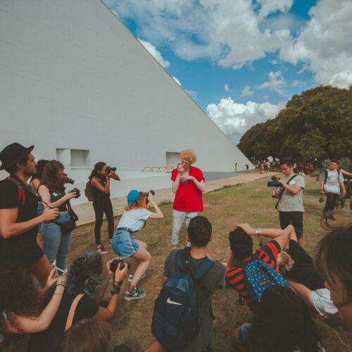 A group of people gathered outdoors, photographing a person in a red shirt, against a geometric backdrop.