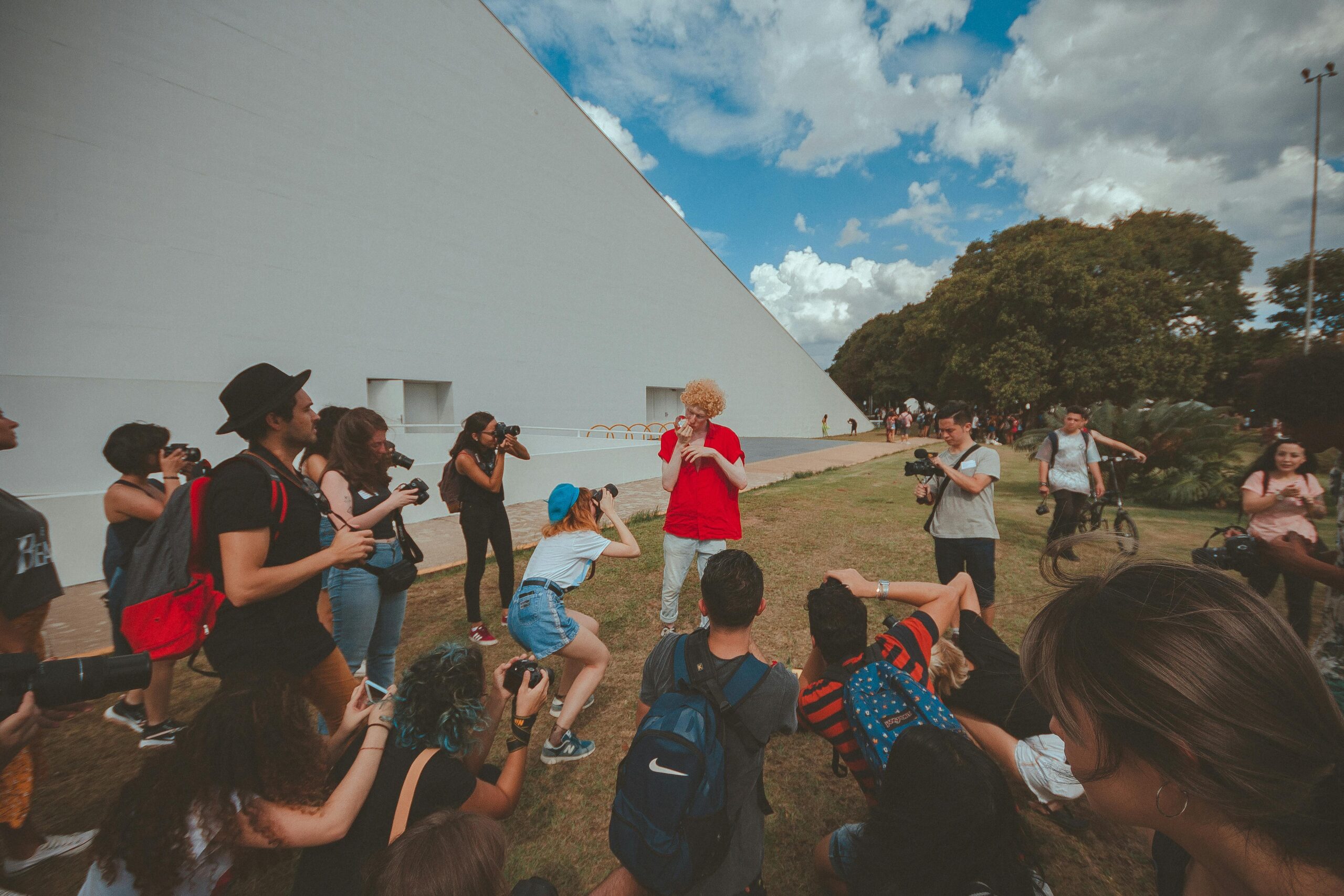 A group of people gathered outdoors, photographing a person in a red shirt, against a geometric backdrop.