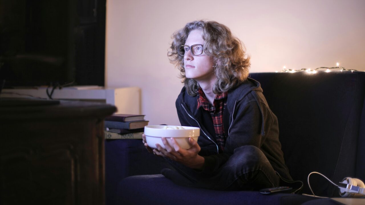 Young man with glasses watching television with focus while holding a snack bowl in a cozy living room setting.