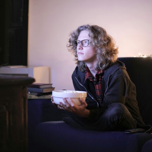 Young man with glasses watching television with focus while holding a snack bowl in a cozy living room setting.