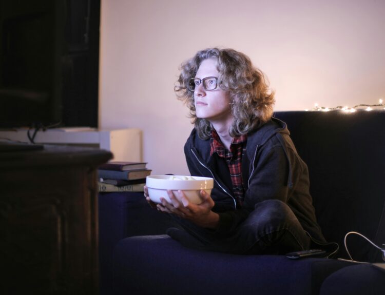 Young man with glasses watching television with focus while holding a snack bowl in a cozy living room setting.