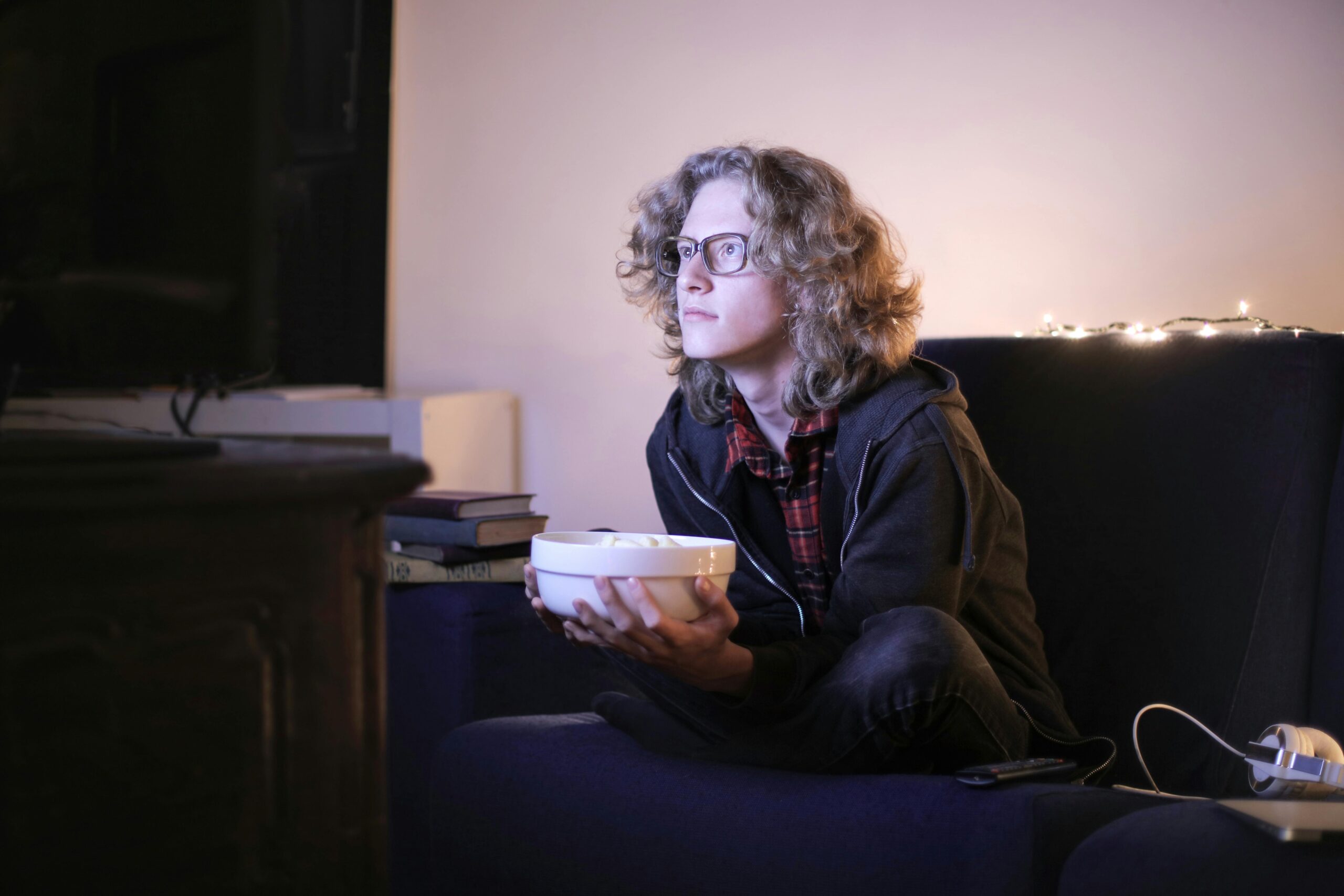 Young man with glasses watching television with focus while holding a snack bowl in a cozy living room setting.