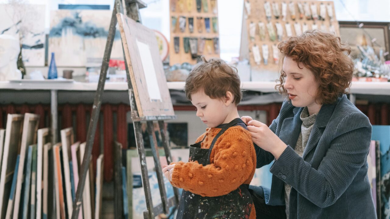 An art teacher helps a young boy with his apron in a vibrant art studio.