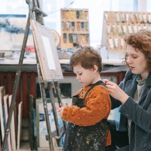 An art teacher helps a young boy with his apron in a vibrant art studio.