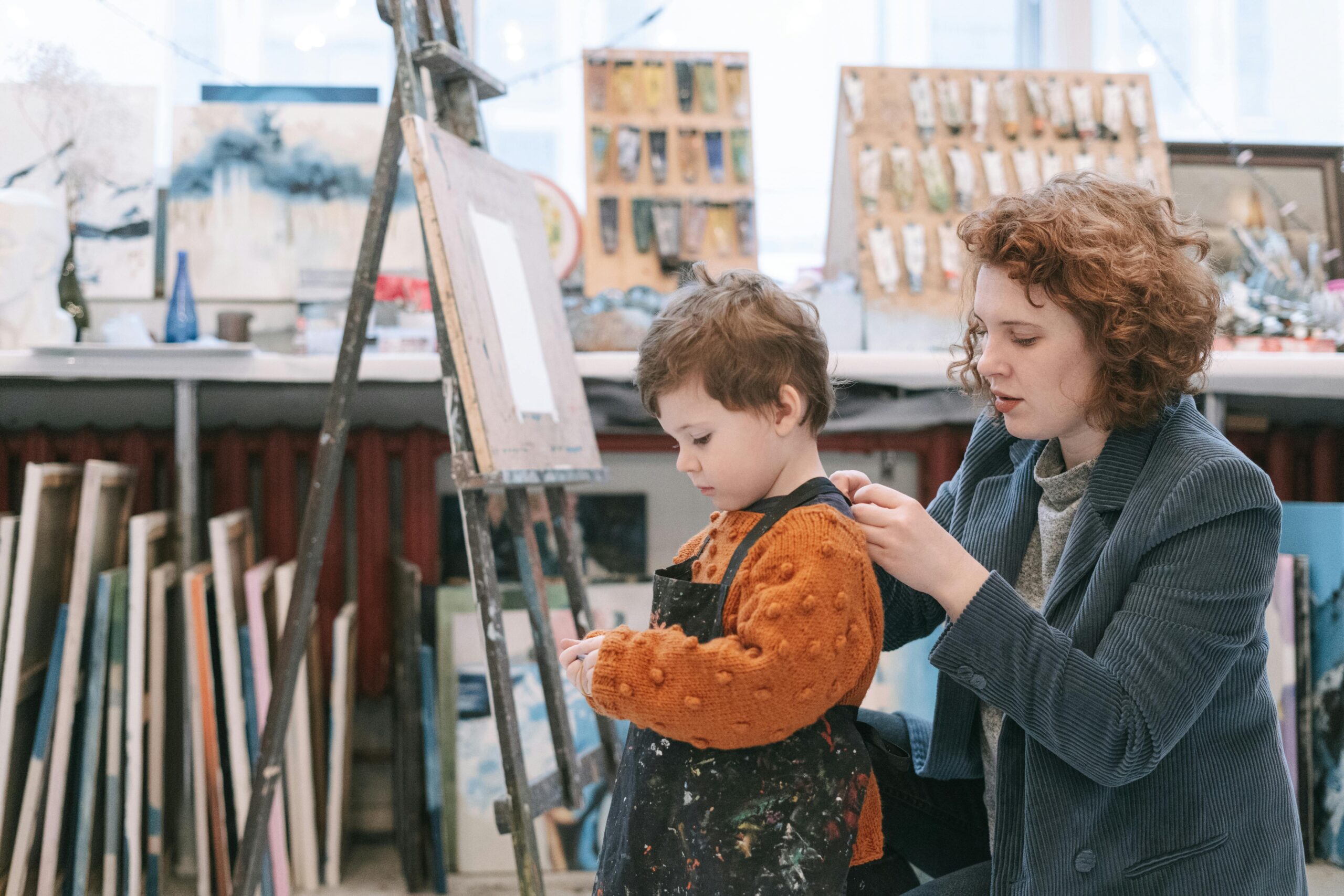 An art teacher helps a young boy with his apron in a vibrant art studio.